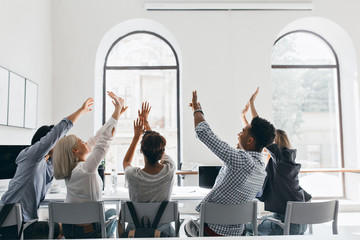 Portrait from back of young people sitting in front of windows in light office. Excited boys and girls which working together celebrating great deal in office.