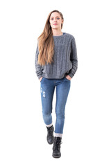 Wall Mural - Cute young pretty woman in sweater and jeans walking towards camera with hands in pockets. Full body isolated on white background. 