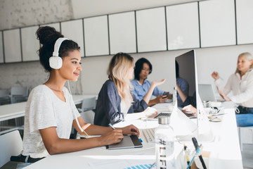 Joyful african web-developer working on new project while listening music in white headphones. Black female designer doing her job in office with talking colleagues on background.