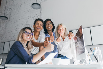 Wall Mural - Asian, african and european specialists having fun in office during coffee break and smiling. Attractive secretary in glasses sitting near computer while her colleagues posing with thumbs up.