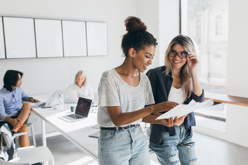 Wall Mural - Gland blonde secretary in glasses looks at african female manager which holding report. Indoor portrait of stylish black woman in jeans spending time in office with asian and european colleagues.