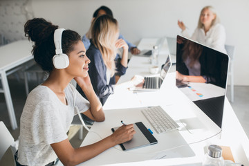 Charming black web-developer listening music in big headphones and using tablet. Indoor portrait of programmers working together in cozy office and drinking coffee.