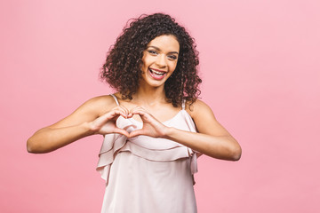 Smiling afro american young girl in dress showing heart with two hands, love sign. Isolated over pink background.