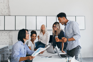 Wall Mural - Pleased asian marketer holding documents and looking at african colleague with smile. Indoor portrait of pretty black woman sitting at the table with laptop beside office friends.