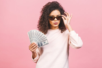 Cool rich girl! Money winner! Surprised beautiful african american woman in casual holding money in sunglasses isolated against pink background.