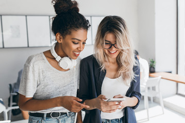 Wall Mural - Gorgeous african female student in casual shirt posing with university mate which texting phone message. Attractive blonde girl working as secretary having fun with international co-worker in office.