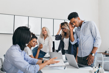 Wall Mural - Confused blonde girl scratches her head while looking at laptop in office. Concentrated african programmer in wristwatch analyses data, discussing something with colleagues.