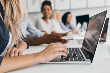 Wall Mural - Blonde woman with elegant hairstyle typing text on keyboard in office. Indoor portrait of international employees with secretary using laptop on foreground.