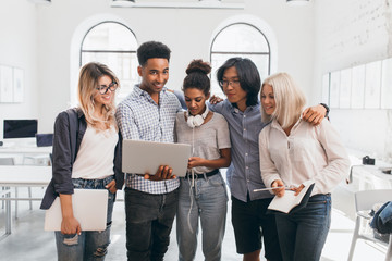 Wall Mural - Blonde female student looks at computer screen and noted something in notebook. Smiling african young man holding laptop, standing in university hall with foreign friends.