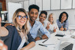 © Look! - African and european girls posing with peace sign while blonde lady making selfie in conference hall. Young african web-developer having fun with colleagues and smiling for photo.