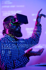 Wall Mural - Young bearded man wearing virtual reality headset gesturing and testing new virtual game while sitting at his desk in dark office