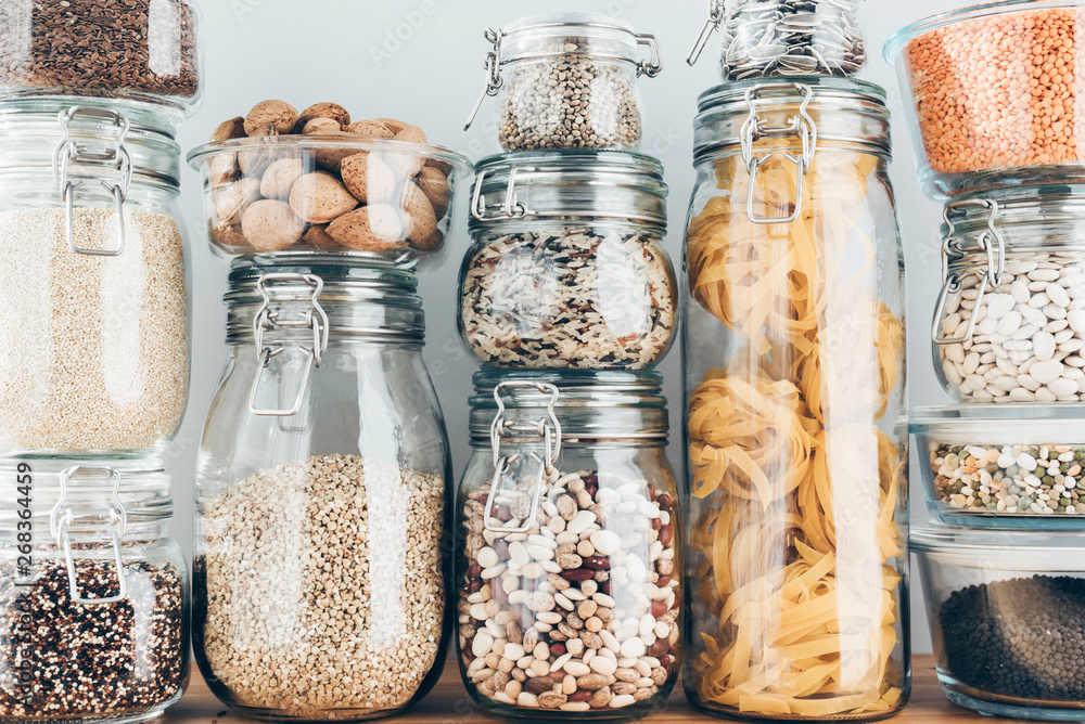 Assortment of uncooked grains, cereals and pasta in glass jars on wooden table. Healthy cooking, clean eating, zero waste concept. Balanced dieting food - obrazy, fototapety, plakaty 