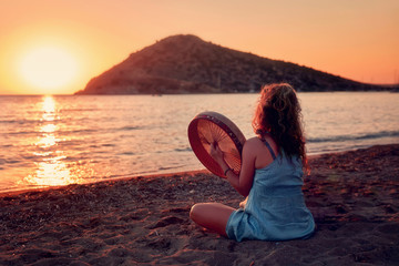 Female hippie sitting on the sands of a beach and playing bendir at  sunset