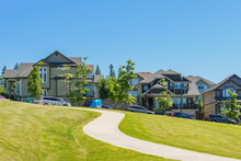 Paved Pathway Over Green Lawn On The Slope In Front Of New Townhouses. Residential Town Homes On Sunny Day With Blue Sky Background