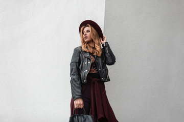 Pretty young woman in a trendy hat in stylish sunglasses in a vintage leather jacket with a backpack with a cape is standing near a white wall in the spring day. Attractive red-haired girl on vacation