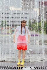 Young wet pretty girl with two braids in yellow boots and with transparent umbrella stands inside of fountain.