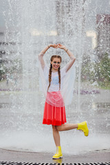 Canvas Print - Young wet pretty girl with two braids in yellow boots and with transparent umbrella stands inside of fountain.