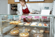 Young Salesman In Red Apron Placing Sweet Pastry In The Refrigerator At The Showcase Of The Confectionery Shop Or Cafe