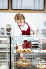 Salesman in red apron putting fresh pie into the refrigerator of the showcase at the modern pastry shop