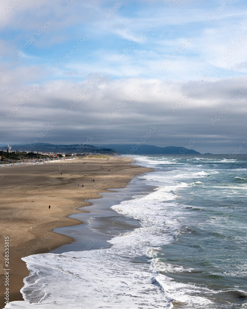 Cloudy afternoon at Ocean Beach in San Francisco. High angle shot looking  south from Cliff House. Mild surf, people enjoying a relatively warm day.  Blue sky above the grey clouds. Stock Photo |, image size:800x1000