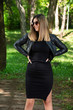 © Emilija - Happy woman in black dress and leather jacket and sunglasses posing in nature on a sunny spring day with hands on hips. Forest in background. Fashion concept. Close up, selective focus