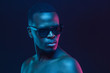 Close-up portrait of african man standing in sunglasses and with naked shoulders in neon light