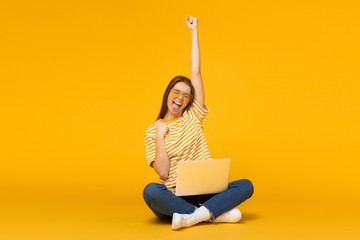 Wall Mural - Studio portrait of joyful girl with laptop computer, sitting on a floor and celebrating, isolated on yellow background