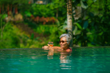 Wall Mural - natural lifestyle portrait of attractive and happy middle aged Asian woman relaxed at tropical resort infinity simming pool with jungle background enjoying a drink relaxed