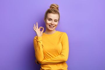 Happy young emotional charming woman showing ok sign with fingers and winking isolated on yellow background, everything is under control.