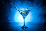 Martini cocktail glass in hand splashing on dark toned smoky background or colorful cocktail in glass with splashes and olives. Party club entertainment. Mixed light. Selective focus