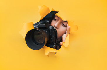 A photographer paparazzi girl with a black DSLR camera looks at what is happening from the ambush with curiosity. Yellow paper, torn hole. Tabloid press. In search of the plot for photo stock.