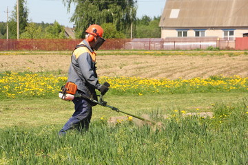 Lawn mower in orange helmet and overalls mows the grass on the village house background on a Sunny summer day - farming, gardening