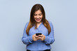 © luismolinero - Young girl over isolated blue wall sending a message with the mobile