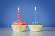 Two Muffins Or Cupcakes With Pink And Blue Burning Candles Against A Blue Background, Birthday Concept With Copy Space