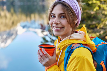 Headshot of smiling woman European female backpacker holds disposable cup of drink, wears yellow coat with hood, poses over blurred lake background with mountains reflection, copy space for text