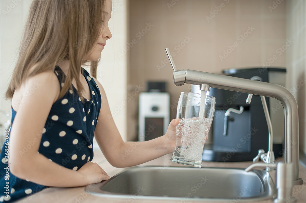 Portrait of a little caucasian girl gaining a glass of tap clean water. Kitchen faucet. Cute curly kid pouring fresh water from filter tap. Indoors. Healthy life concept - obrazy, fototapety, plakaty 