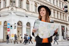 Young Positive Tourist Holds Travel Paper Map, Reads Route Of Trip, Goes Sightseeing, Searches Destination, Gets To Right Place Wears Black Headgear And Leather Jacket Focused Away With Pleasant Smile