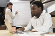 © Anatoliy Karlyuk - Confident concentrated African man using portable electronic gadget at office desk while his female assistant in background working on presentation, standing at flip chart, reporting about project