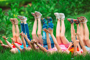 Wall Mural - Children lying on green grass in park on a summer day with their legs lifted up to the sky.