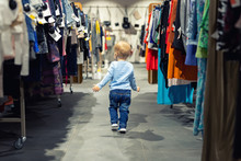 Cute Caucasian Blond Toddler Boy Walking Alone At Clothes Retail Store Between Rack With Hangers. Baby Discovers Adult Shopping World. Baby Get Lost At Big Hypermarket Shopping Mall