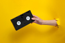 Female Hand Holds Video Cassette Through Torn Yellow Paper Background. Minimalistic Retro Concept
