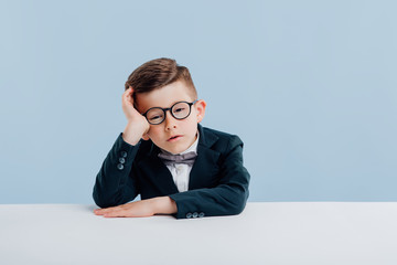 Wall Mural - education. little schoolboy in glasses holding hands in his head, dressed in school uniform, looking at camera, sitting at the white table, isolated on blue background, copy space, school concept