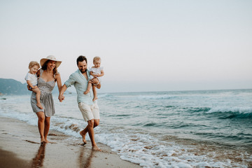 a family with two toddler children walking on beach on summer holiday at sunset.