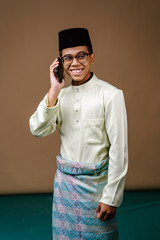 Wall Mural - Studio portrait of a young and intelligent looking Malay Asian man in a baju melayu, sampin, songkok hat and spectacles. He is smiling as he uses his smartphone. He is dressed for Ramadan. 