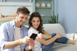 © ty - Young couple watching online content in a smart phone sitting on a sofa at home in the living room.