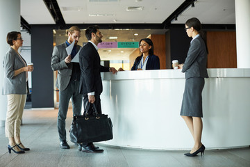 Handsome mixed race businessman with travel bag standing at registration counter and talking to receptionist while checking in to hotel