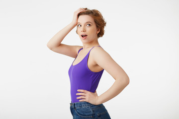 Poster - Portrait of hocked attractive short-haired girl, wearing a purple jersey, looking away in surprise with wide open mouth and eyes, straightens hair with his hand, isolated over white wall.