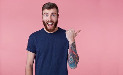 Wall Mural - Young happy red-bearded young guy , with wide open mouth in surprise, saw something wrong, wants to draw your attention pointing finger to copy space at the right side isolated over pink background.