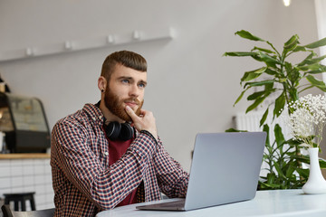 Wall Mural - Young attractive ginger bearded man sits at a table in a cafe and works on a laptop, wearing in basic clothes, thoughtfully looking away, trying to come up with a solution.