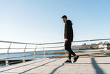 Fototapeta Młodzieżowe - Photo of joyful guy 20s walking along wooden boardwalk by seaside after morning workout